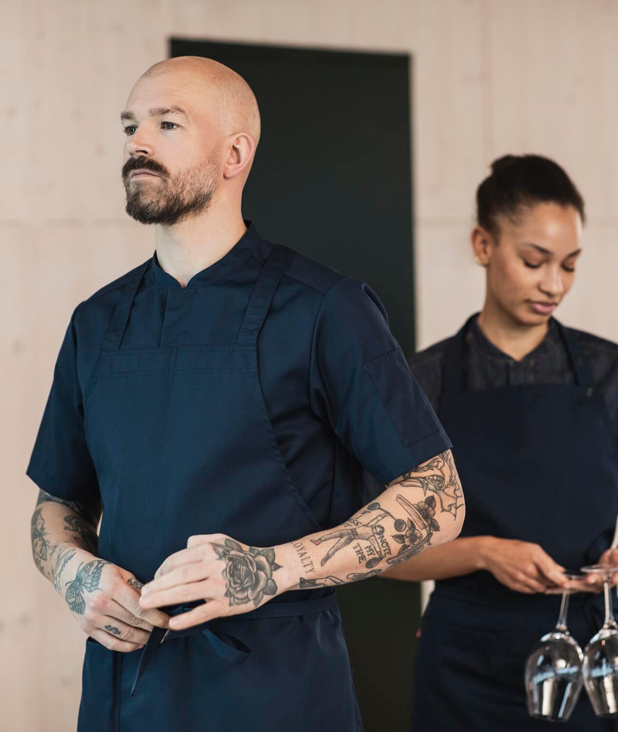 Två professionella kockar klädda i Segers senaste mörkblå kockrock modell 1097 och förkläde modell 4579 arbetar samfokuserat i ett kök med modern design, framhäver tatuerade armar som symbol för hantverksskicklighet.