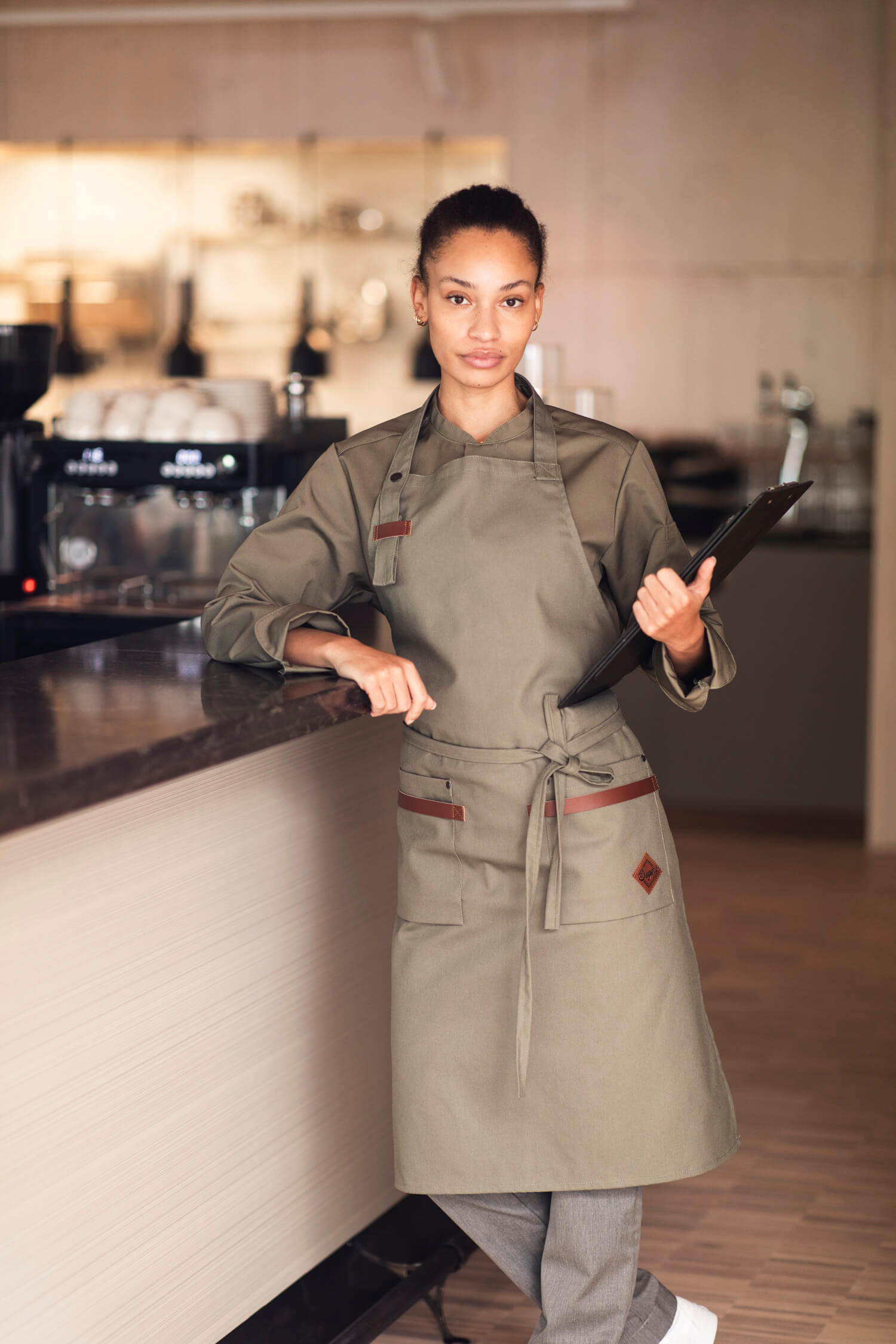 Professionell kvinnlig kock poserar säkert i restaurangmiljö, iklädd Segers nyaste kockskjorta modell 1099 och förkläde modell 4093 i en elegant olivgrön färg, redo för kulinariska utmaningar.