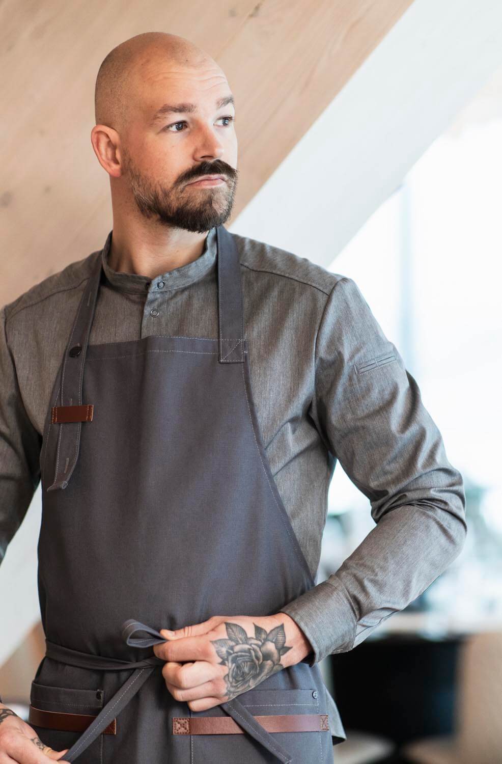 Professionell kock i Segers nya kollektion, med snygg kockskjorta modell 1109 och stilrent förkläde modell 4093, visar upp en tatuering på underarmen i en modern restaurangmiljö.