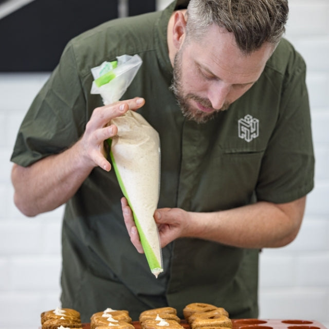 Man som spritsar fyllning till bullar klädd i olivgrön kockskjorta.