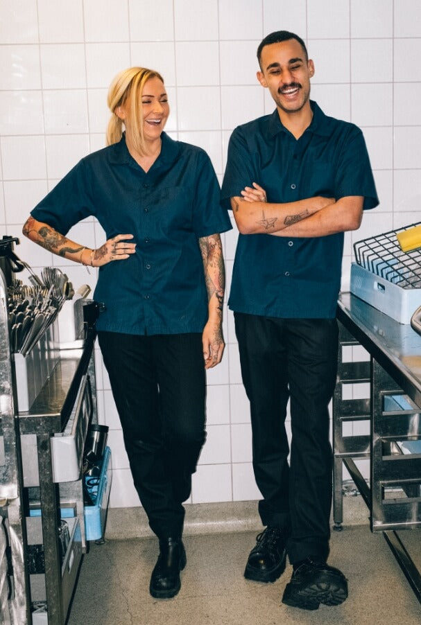 Två personer i restaurangkök som bär mörka arbetskläder. De står bredvid varandra och skrattar, med köksutrustning och rostfria ytor i bakgrunden.