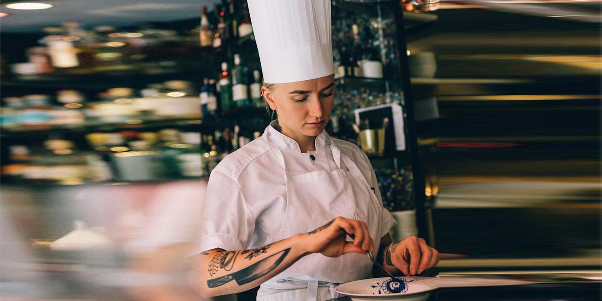 En professionell kvinnlig kock iklädd en Segers kockrock modell 1024 koncentrerar sig på att finslipa en anrättning i ett livligt restaurangkök, hennes tatueringar tillför en personlig touch till den klassiska vita kockuniformen.