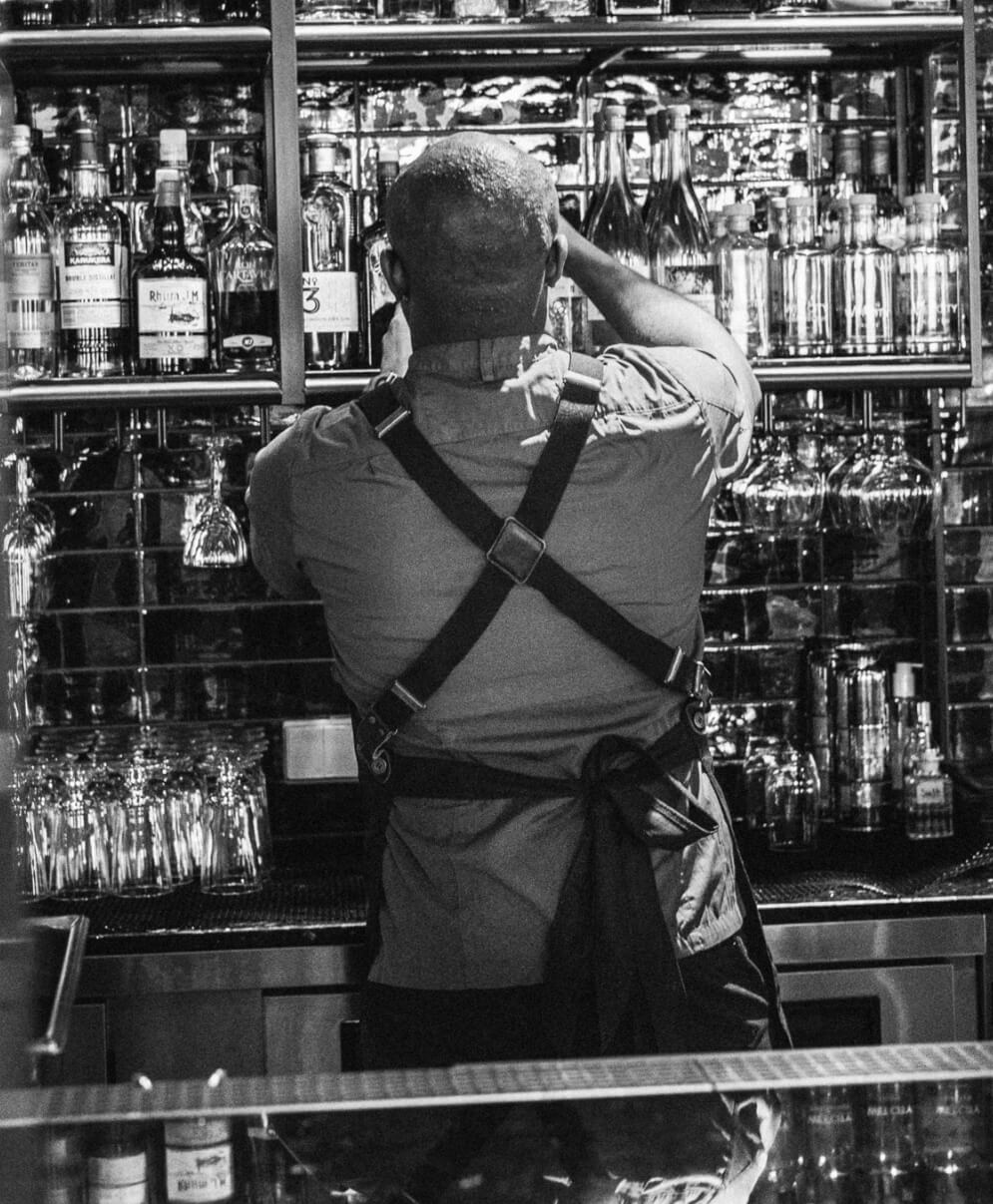 Bartender sedd bakifrån bär förkläde med korsade axelremmar vid barhylla.