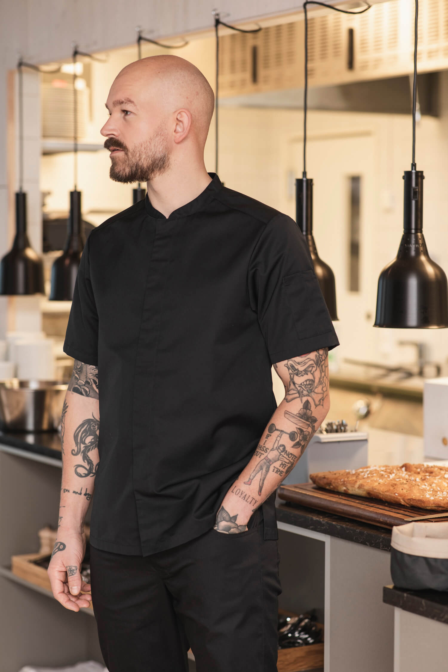  Kock med Segers nya kockrock modell 1097 står i ett modernt kök med armar prydda av tatueringar.